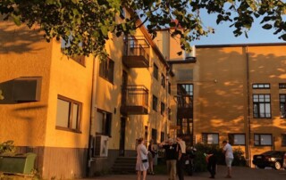 Oranssinkeltainen iso rakennus, jonka edellä eri ikäisiä ihmisiä keskustelemassa. Kuvan yläosassa puiden oksia. Puiden varjot kuvastuvat myös rakennuksen seinässä.