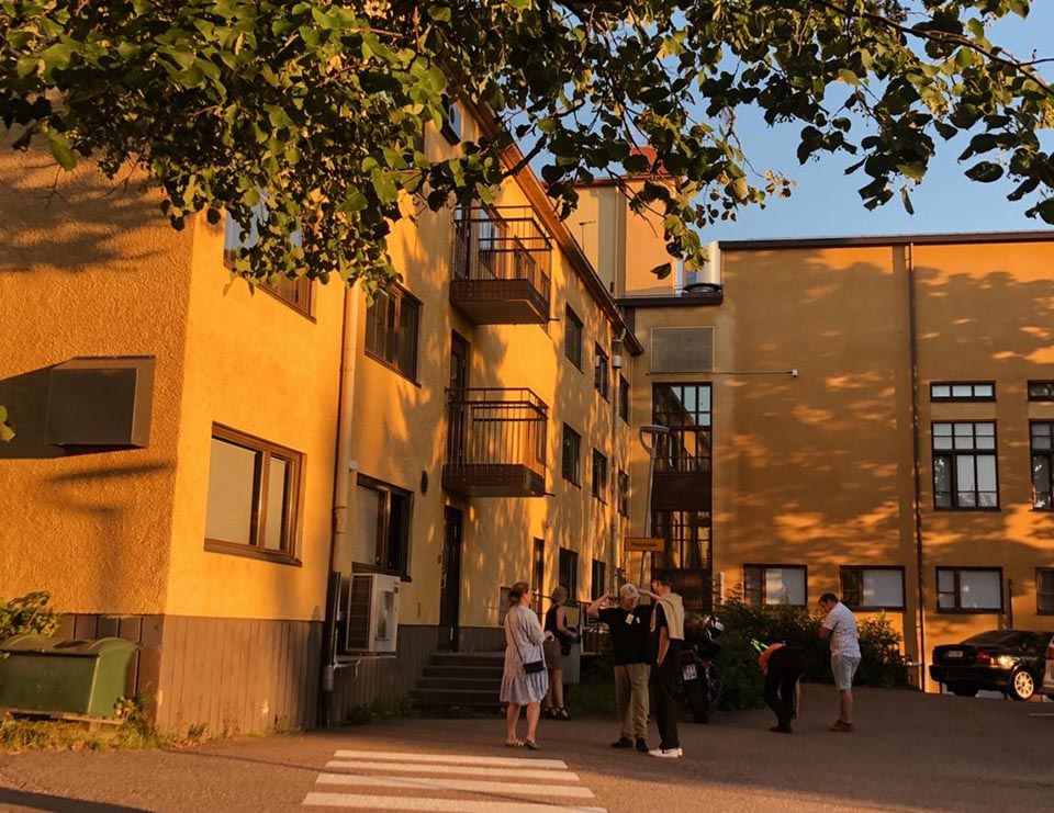 Oranssinkeltainen iso rakennus, jonka edellä eri ikäisiä ihmisiä keskustelemassa. Kuvan yläosassa puiden oksia. Puiden varjot kuvastuvat myös rakennuksen seinässä.